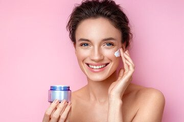 Beautiful woman applying moisturizer cream on her face. Photo of woman with perfect skin on pink background. Beauty and Skin care concept