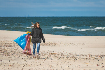 Beautiful blond girl collecting plastic trash rubbish garbage on the sand beach and putting it into big plastic bags for recycle. Cleaning and recycling concept, campaign to clean volunteer concept - obrazy, fototapety, plakaty