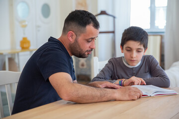 Concentrated father and son sitting at table and reading book. Bearded dad trying to help his son with task. Focused Caucasian boy looking at book. Education, family time concept