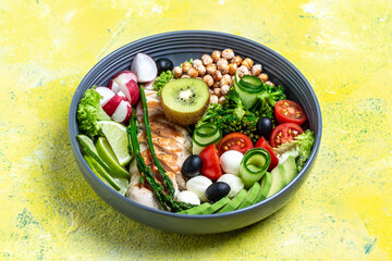 Healthy salad bowl with avocado, asparagus, chickpeas, broccoli, radish, chicken, cucumber, tomatoes, olives, mozzarella on yellow background. top view. Food and health