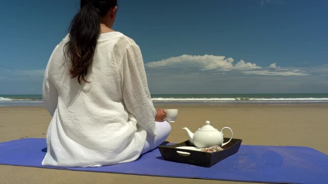 wellness and relaxation concept - back shot of healthy and beautiful asian woman in white sitting on exercise mat with cup of tea and practice yoga lotus pose for meditation on the tropical beach