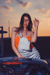 Sexy young female in white overall and orange t-shirt is posing outdoors standing in car cabriolet....