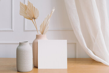 Square white paper mockup and modern ceramic vases with dried palm leaves. Stylish bright living...