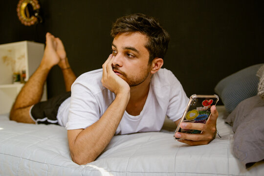 Joven gay descansando en su cama mientras utiliza su teléfono celular 