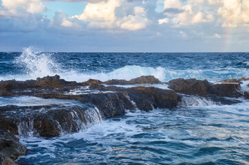 Waves splashing and running over rocks in the ocean with a small rainbow in the sky on a fall evening in Qawra, Malta.