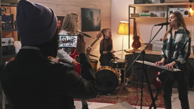 Medium slowmo of girl in cool leather jacket and beanie hat recording video on smartphone of all-girl rock band singing and playing musical instruments in retro-style studio