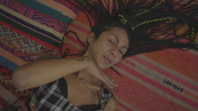 Mujer transgénero con trenzas y tatuajes haciendo gestos acostada sobre la alfombra de su habitación 