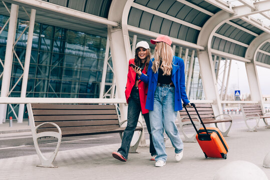 Two happy girls using smartphone checking flight or online check-in at airport together, with luggage. Air travel, summer holiday