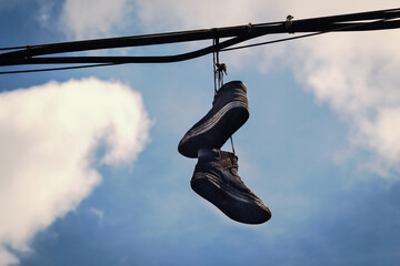 Two dirty old sneakers hanging on wires in blue sky with clouds - obrazy, fototapety, plakaty