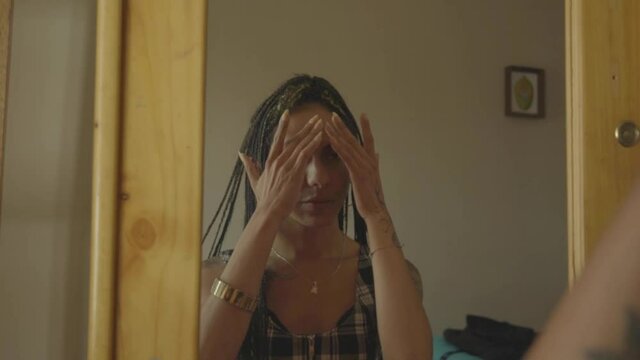 Mujer transg&eacute;nero con trenzas apreciando su rostro frente al espejo de su habitaci&oacute;n 