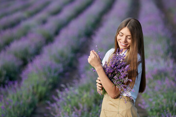 Portrait of pretty young smiling female wearing farm apron collecting lavender harvest in summer. Happy girl posing with bouquet near patches in endless lavender field and looking aside.
