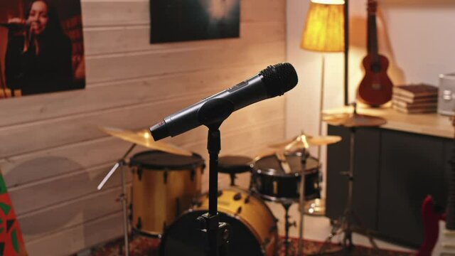 Slowmo closeup of black microphone on mic stand and drum set and acoustic and electronic guitars in retro-style music studio