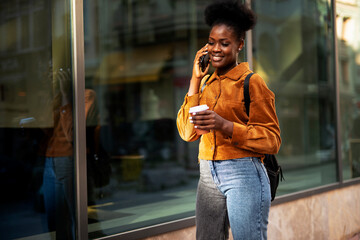 Beautiful young woman drinking coffee while walking around town. Young african woman using the phone..