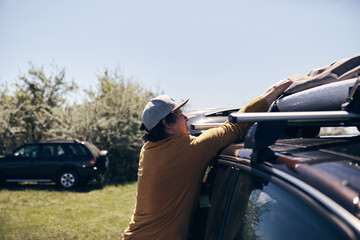 Windsurfer and camper packing and unpacking from a car's roof rack in nature.