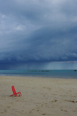 Fototapeta na wymiar A small red chair on the deserted beach while a thunderstorm is coming