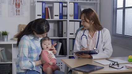 asian mother holding baby talking to pediatrician in hospital room. female doctor holding clipboard...
