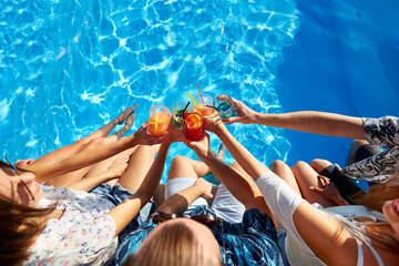 Top view of of friends clinking glasses with fresh colorful cocktails sitting by swimming pool on...