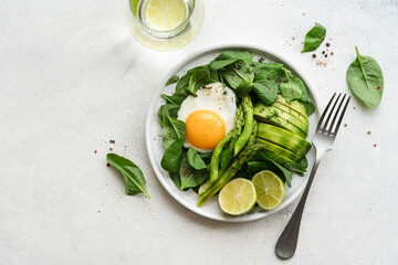 Healthy green breakfast with fried egg. Asparagus, spinach, avocado and egg in plate on white stone background