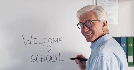 Aged professor writing welcome to school on white board and smiling at camera