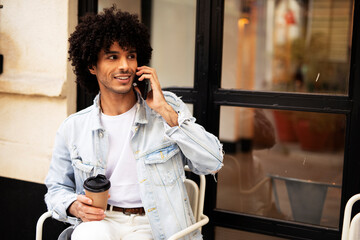 Young man drinking coffee outdoors. Handsome man with curly hair talking to the phone.