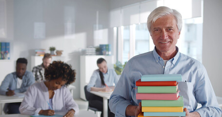 Fototapeta premium Portrait of aged male teacher holding stack of books and smiling at camera in class