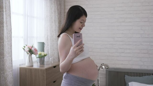 happy pregnant mother facing camera and holding a mobile phone is taking a mirror selfie at home with white brick wall in the background.
