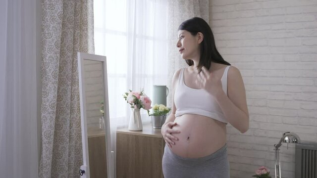 cheerful mom expecting her baby is tucking her hair and putting hand on the waist while watching herself in the mirror at home.