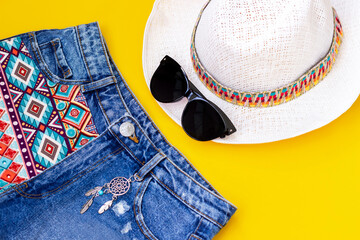 Stylish short denim shorts with a boho pattern and a dream catcher, black sunglasses and white straw hat isolated on a bright color yellow background. Top view. Summer vacation concept	