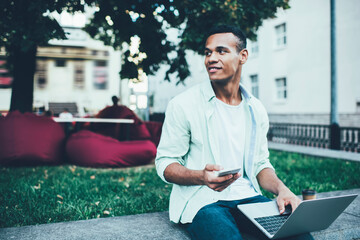 Cheerful man working on laptop and messaging on smartphone in park