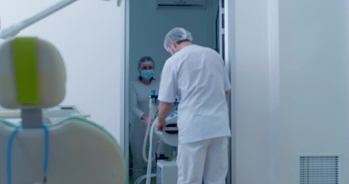The female and male medical workers are wearing disposable masks and hats are rolling a table with an anesthesia workstation into the room.