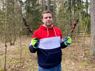 A man holds skewers with toasted pork skewers in his hands. Outdoor recreation. Fry a delicious shish kebab in the forest