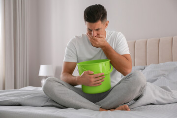 Man with bucket suffering from nausea on bed at home. Food poisoning - obrazy, fototapety, plakaty