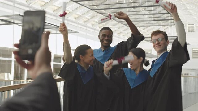 Back view of unrecognizable man making photo of four young multiethnic graduates wearing university graduate gowns and holding diplomas using smartphone camera