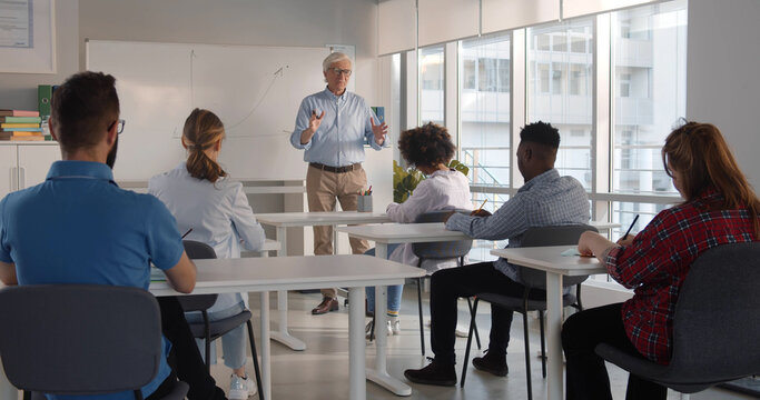 Senior Teacher Explaining Diagram Written On Whiteboard While Diverse College Students Sitting At Desk