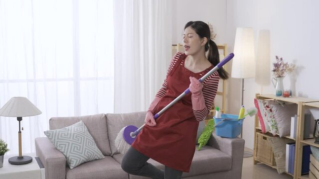 energetic asian wife is partying alone in the living room at home by imagining herself a rocker chanting and playing the bass guitar with a swabber.
