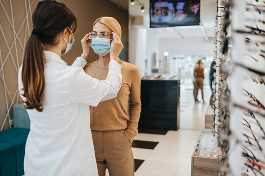 Beautiful woman choosing eyeglasses frame in modern optical store. Female seller specialist helps her to make right decision. They are wearing protective face mask against virus pandemic.