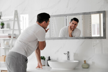 Fototapeta premium Handsome man touching his smooth face after shaving near mirror in bathroom