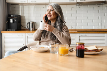 Mature smiling woman talking on cellphone while having breakfast