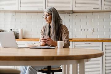 Mature grey woman working with laptop and cellphone in kitchen