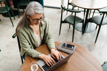 Mature grey woman using mobile phone while working with laptop