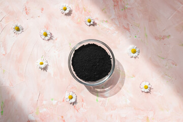 Black activated charcoal powder in a glass bowl on pastel pink floral background. Ingredient for beauty, cosmetics, spa treatment, detox concept, copy space. Sunlight and shadows trendy shot