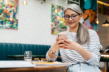 Mature grey woman smiling and using cellphone while sitting in cafe