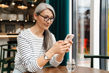 Mature grey woman using mobile phone while sitting in cafe indoors