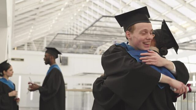 Medium shot of young Caucasian male graduate hugging his mixed-race female friend wearing university graduate gowns and caps. Then they talking and showing diplomas to each other