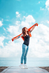 A young happy woman reaches up with her hand. Vertical orientation. Copy space. In the background, the blue sky with clouds and the ocean. Concept of freedom and carefree