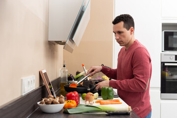 Man wearing an apron cooking at home in a modern kitchen vegetarian and vegan food looking at a tablet. Healthy recipe.