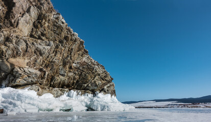 Fototapeta na wymiar A granite rock, devoid of vegetation, against the background of a blue sky and a frozen lake. Deep cracks on the stones. On the base - ice splashes, icicles. Winter sunny day. Baikal