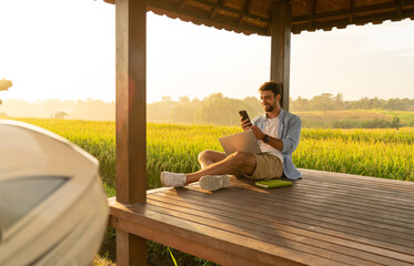 Positive young guy chilling in field gazebo with smartphone and laptop