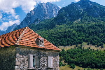 Fototapeta na wymiar View of traditional Albanian stone house with orange slate roof and wooden shutters in mountain village. Lush foliage and blue sky.
