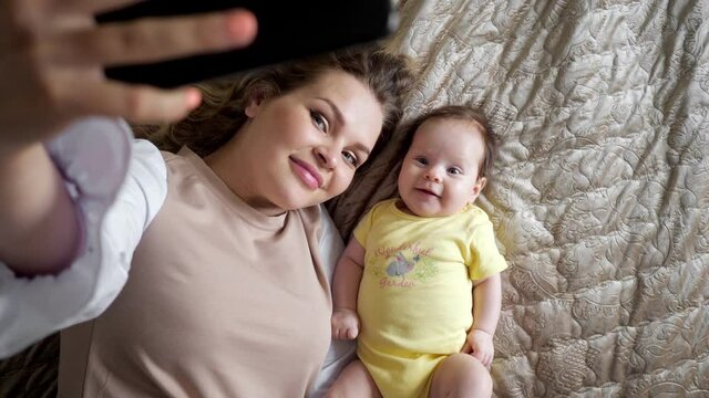 Jolly young mother with plump lips takes picture of smiling baby daughter in yellow with smartphone on bed in apartment room closeup